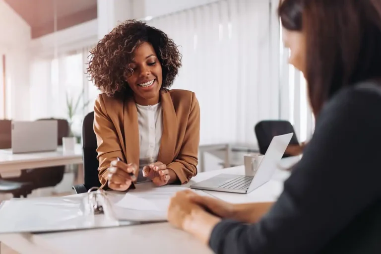 In a professional office environment, two engaged team members focus on La Crosse SEO strategies. One, with curly hair and a tan blazer, gestures energetically towards the documents on the table. The other attentively reviews them, aided by an open laptop displaying additional information.