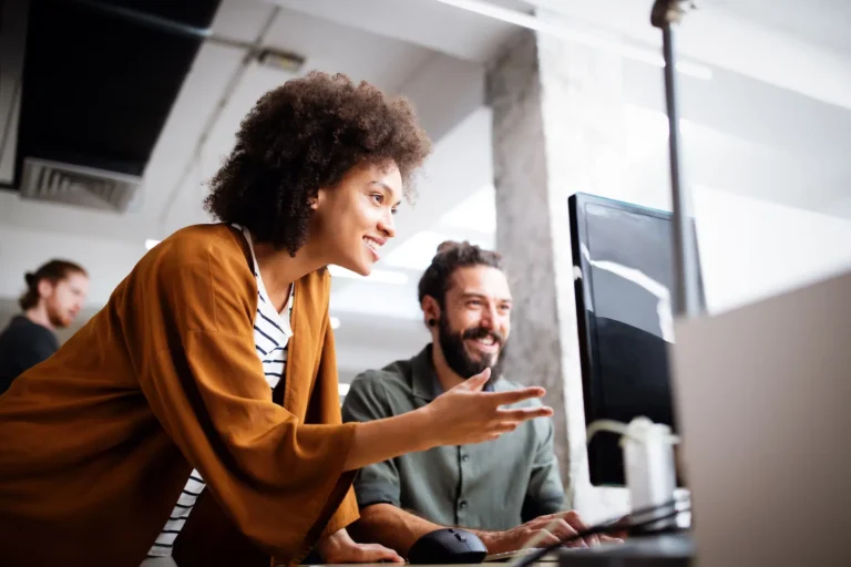 In a modern office, two professionals engage in a dynamic discussion on Mankato SEO strategies. The woman with curly hair points emphatically at the computer screen, while the bearded man beside her nods appreciatively. In the background, colleagues continue their work amidst softened chatter.