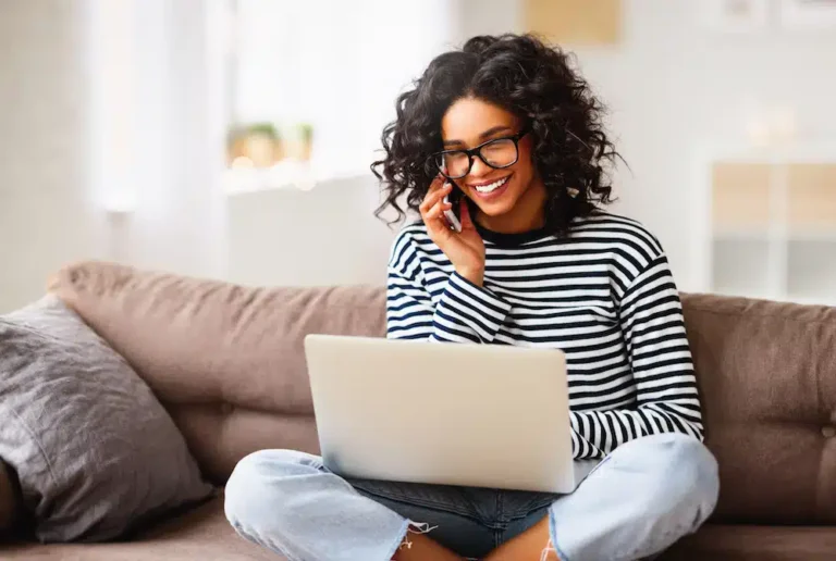 A curly-haired woman in glasses sits cross-legged on a couch, engaging in a discussion about Minneapolis PPC strategies. Using her laptop, she exudes confidence and expertise. Dressed in a striped shirt and jeans, she operates from a bright, welcoming living room setting.