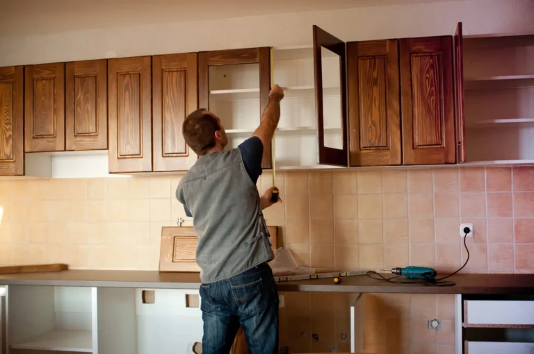 Expertly assembling wooden kitchen cabinets for a remodeling project, a skilled professional secures a shelf inside an open cabinet. The kitchen boasts brown cabinets, tiled backsplash, and new countertops. Tools are strategically placed around the area, emphasizing the transformational process involved in this renovation.