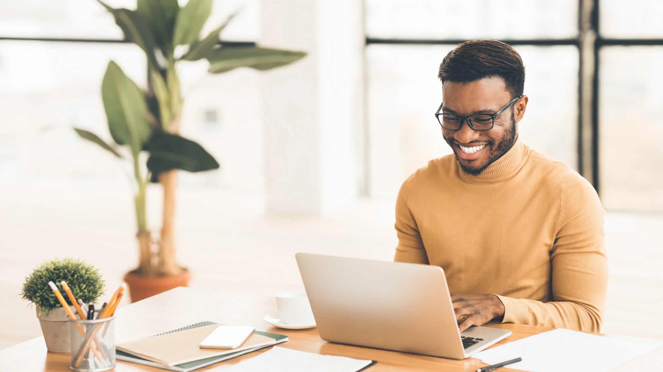 A man in glasses and a brown sweater sits at a desk, focused on developing San Antonio SEO strategies on his laptop. The space is bright and organized, featuring a plant, notebooks, a white cup, and a smartphone with natural light streaming through large windows.