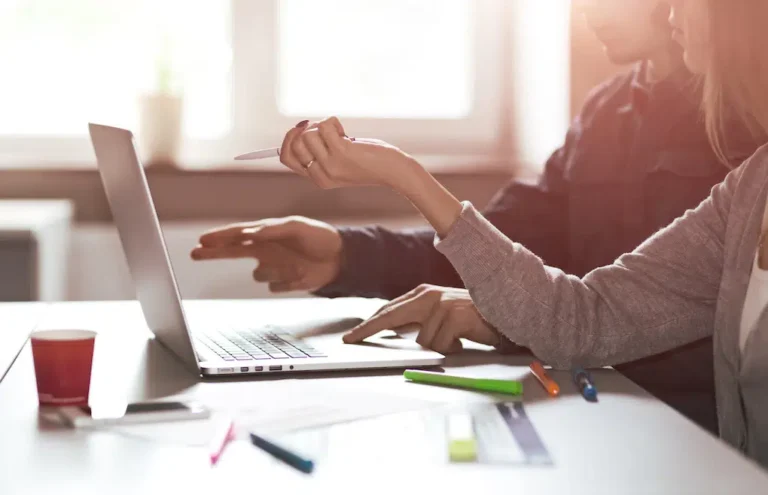 In a sunlit San Antonio office, two individuals engage in an SEO brainstorming session. One expertly navigates the laptop touchpad while the other highlights key points on the screen with a pen. Surrounded by vibrant stationery, they collaboratively develop innovative strategies for online growth.