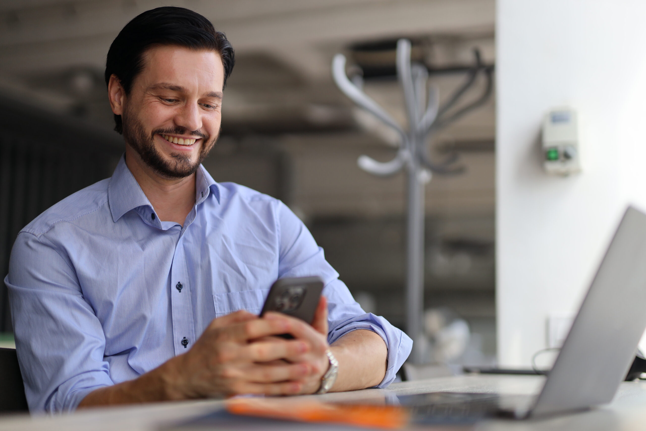 A man in a blue shirt smiles as he reviews Des Moines SEO updates on his phone, seated at his desk with a laptop in an office setting.