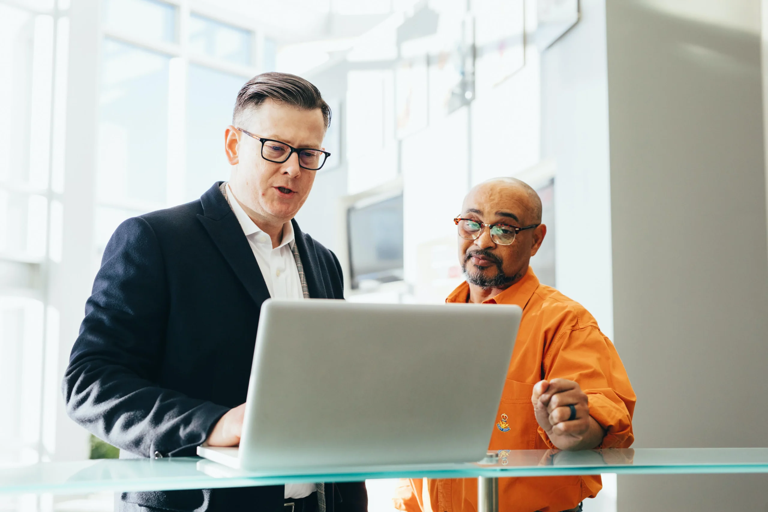 In a modern, well-lit office, two professionals engage in a focused discussion about La Crosse SEO at a glass table. One, dressed in a dark suit and glasses, collaborates with his colleague wearing an orange shirt and glasses as they examine the information on their laptop.
