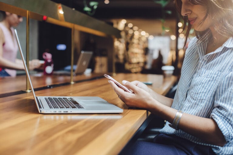 In a modern, well-lit cafe frequented by Columbus SEO experts, a woman sits at a wooden counter. She is focused on her smartphone, with an open laptop in front of her. Another individual nearby is equally engaged with their device.