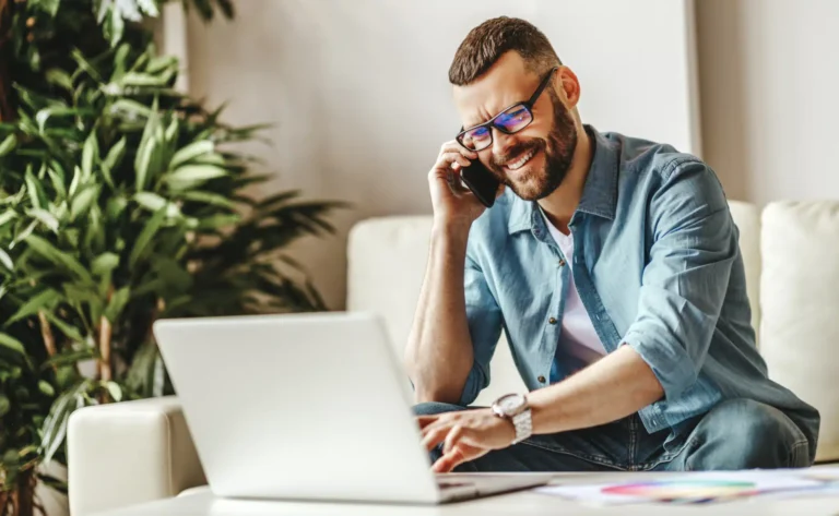 A man in a light denim shirt and jeans, smiling while discussing Wichita web design on the phone, sits in a bright room adorned with plants, utilizing his laptop.