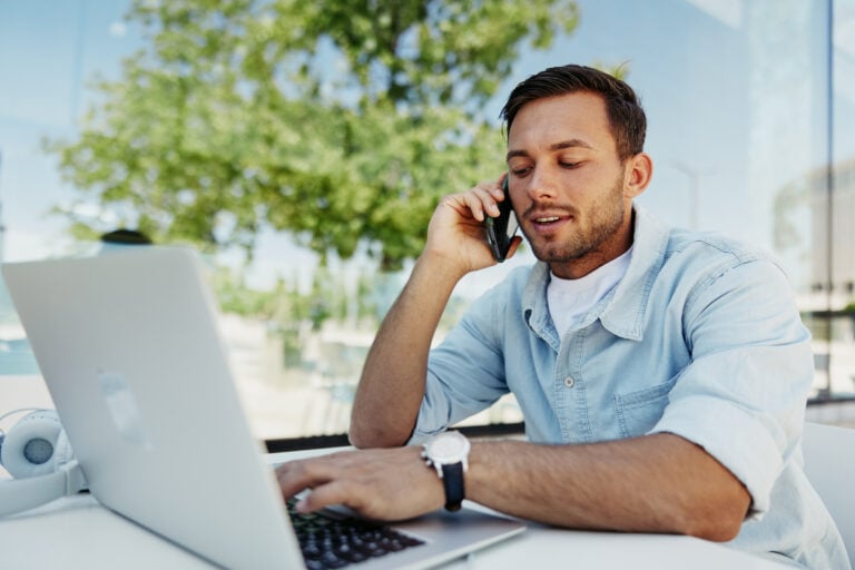 A man in a light denim shirt sits at an outdoor table, simultaneously managing a phone call and working on his laptop under a clear, sunny sky. Behind him, the reflection of a tree and glass building highlights the dynamic urban landscape of Wichita SEO.