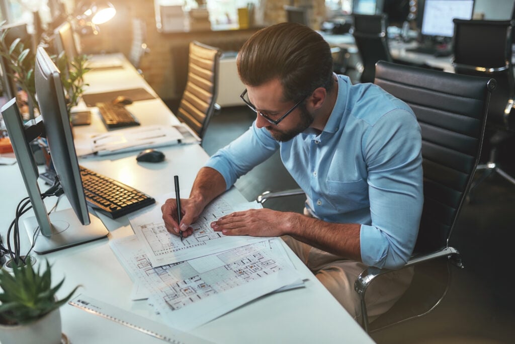 Professional architect at desk reviews blueprints and brainstorms firm names; monitors, keyboard, and plants are visible.