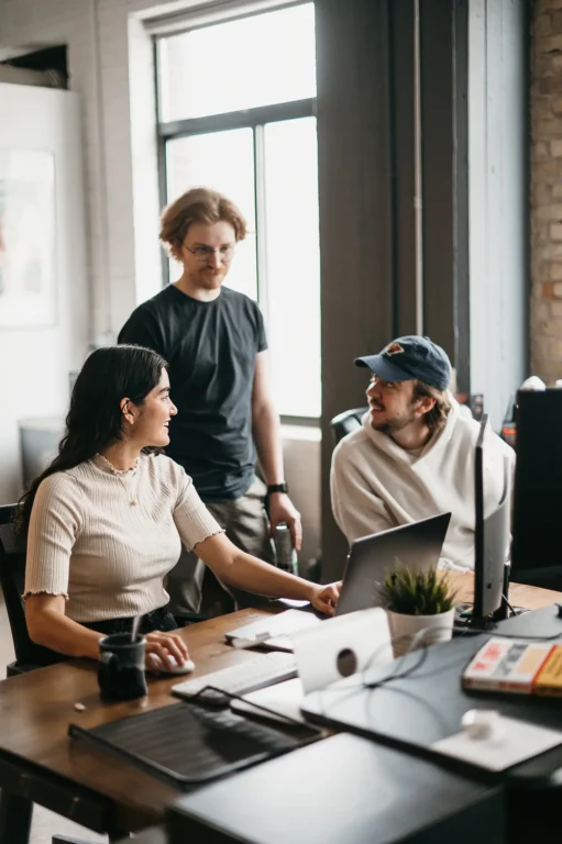 In a relaxed office setting, three colleagues gather around a desk. A woman sits at her laptop, engaging warmly with two standing colleagues. One leans casually on the desk, sharing SEO insights with a friendly demeanor.