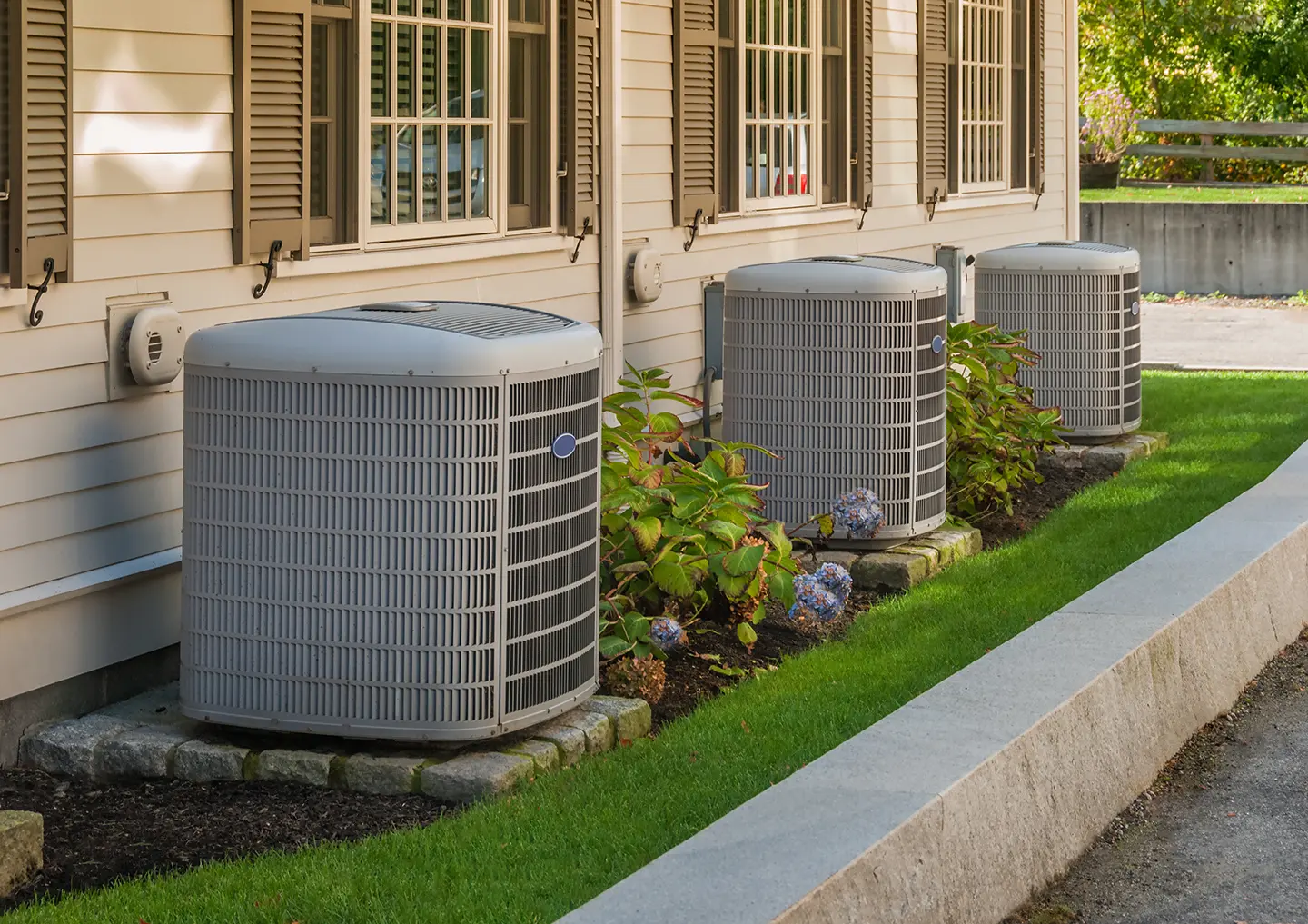Three outdoor air conditioning units, expertly serviced by HVAC LSA Services, are strategically positioned beside a house with beige siding and shuttered windows. Resting on concrete slabs, these units are surrounded by neatly trimmed grass and small shrubs. A well-maintained paved path runs alongside the setup.