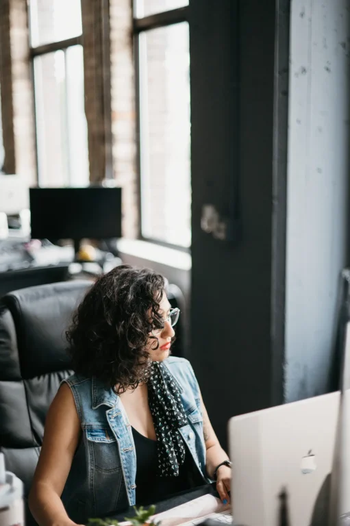 A professional sporting curly hair and sunglasses works diligently at an iMac, researching LSA services. They are dressed in a denim jacket and scarf, situated in a well-lit office with expansive windows and modern equipment.