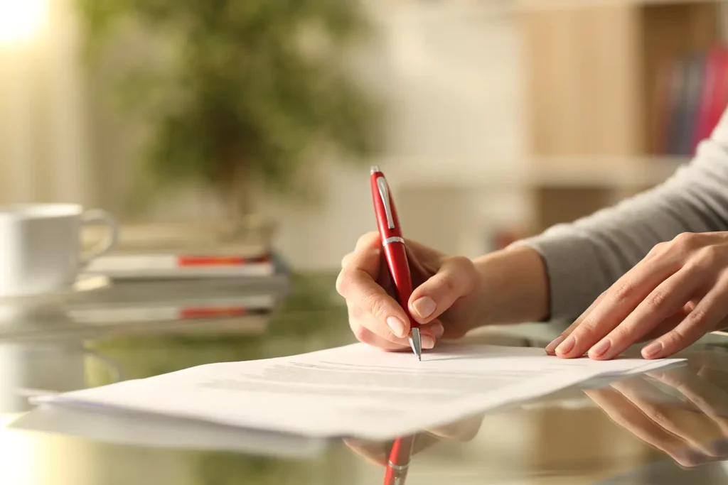 On Hook Agency's website, a professional setting is depicted: An individual is signing a roofing contract using a red pen on a glass table. In the background, blurred elements include a stack of books, a white coffee cup, and a green plant. The well-lit scene emphasizes the importance of the task at hand, fostering an atmosphere of professionalism and focus.