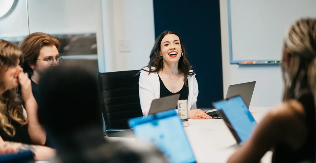 A team gathers around a conference table, focused on analyzing the new website's results. A leader in a white shirt, smiling confidently, introduces an innovative new strategy. The room is brightly lit, with a whiteboard and markers in the background.