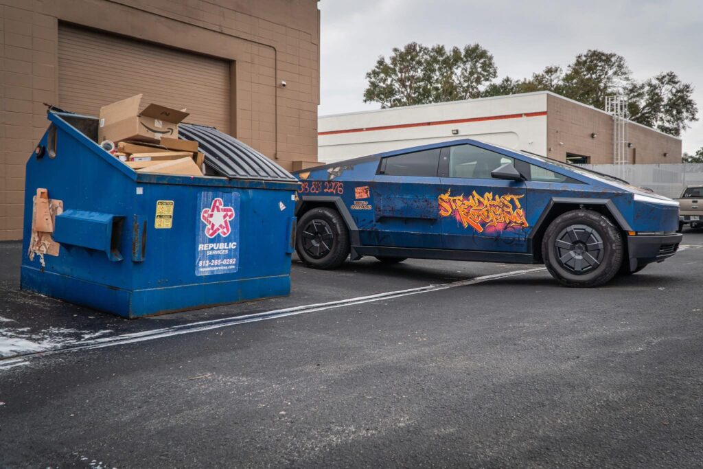 On Hook Agency's website: A blue vehicle adorned with striking graffiti and innovative designs is parked next to a blue dumpster full of cardboard boxes. The scene is set in an industrial area, surrounded by brown and white buildings with trees in the background.