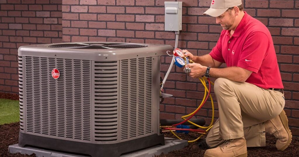 A technician in a red shirt and beige pants expertly inspects an outdoor air conditioning unit. Utilizing precision gauges, he ensures optimal performance and efficiency alongside a brick wall.