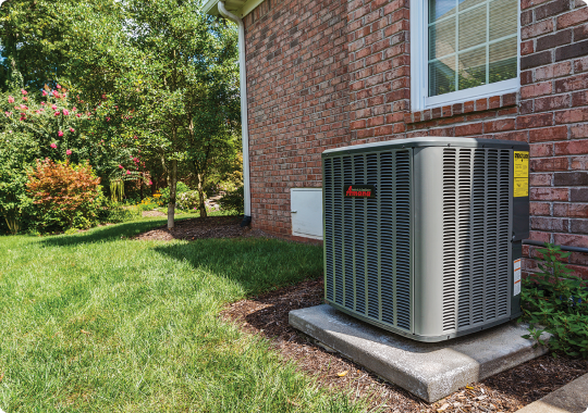 An industry-leading outdoor air conditioning unit is installed on a concrete slab adjacent to a brick home. The area is enhanced by a manicured lawn, a small garden bed, and strategic shrubbery. Sunlight streaming through the trees creates an inviting play of shadows across the landscape.