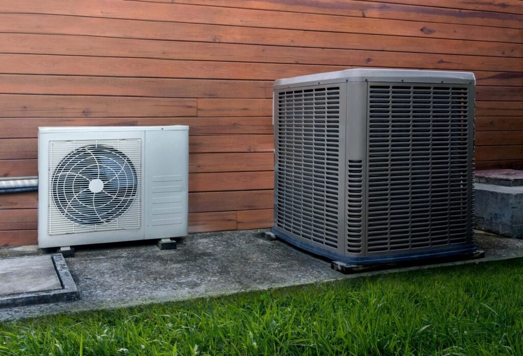Experience the perfect harmony of form and function with our top-tier HVAC solutions. On display are two outdoor air conditioning units from leading brands, strategically placed on a concrete slab for stability. The sleek, white unit complements the robust, gray one, while lush grass surrounds them, enhancing their aesthetic appeal. Choose quality craftsmanship for your climate control needs at Hook Agency.