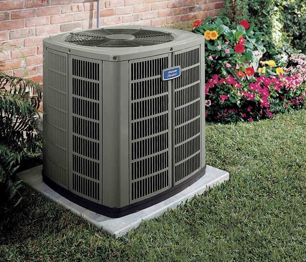 A high-quality outdoor air conditioning unit from a leading HVAC brand is securely installed on a concrete slab adjacent to a brick wall. The unit is surrounded by lush green grass, vibrant flower beds, and blooming plants.