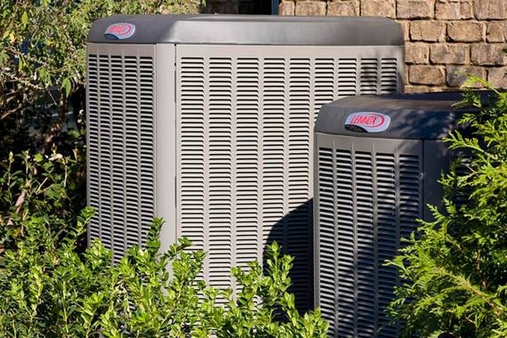 On the Hook Agency website, two prominent gray outdoor air conditioning units from leading HVAC brands are showcased against a brick wall. Surrounded by lush green bushes and trees, each unit prominently displays the manufacturer's logo.