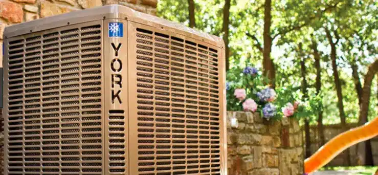 On a sunny day, a York air conditioning unit stands outdoors, framed by a stone wall and vibrant flowers. Surrounded by lush green trees, it embodies reliability and serenity in one exceptional HVAC system.