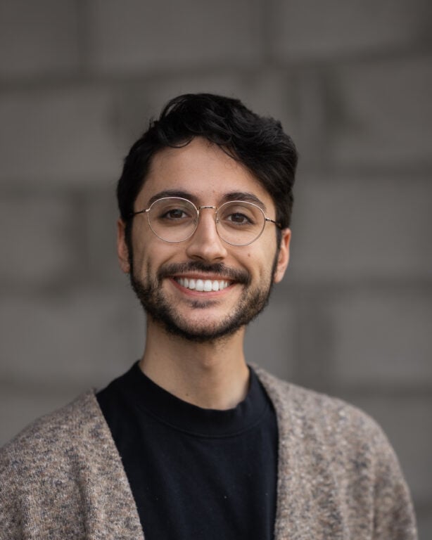 Professional team member with short dark hair, glasses, and beard; photographed against a blurred gray background for Hook Agency.