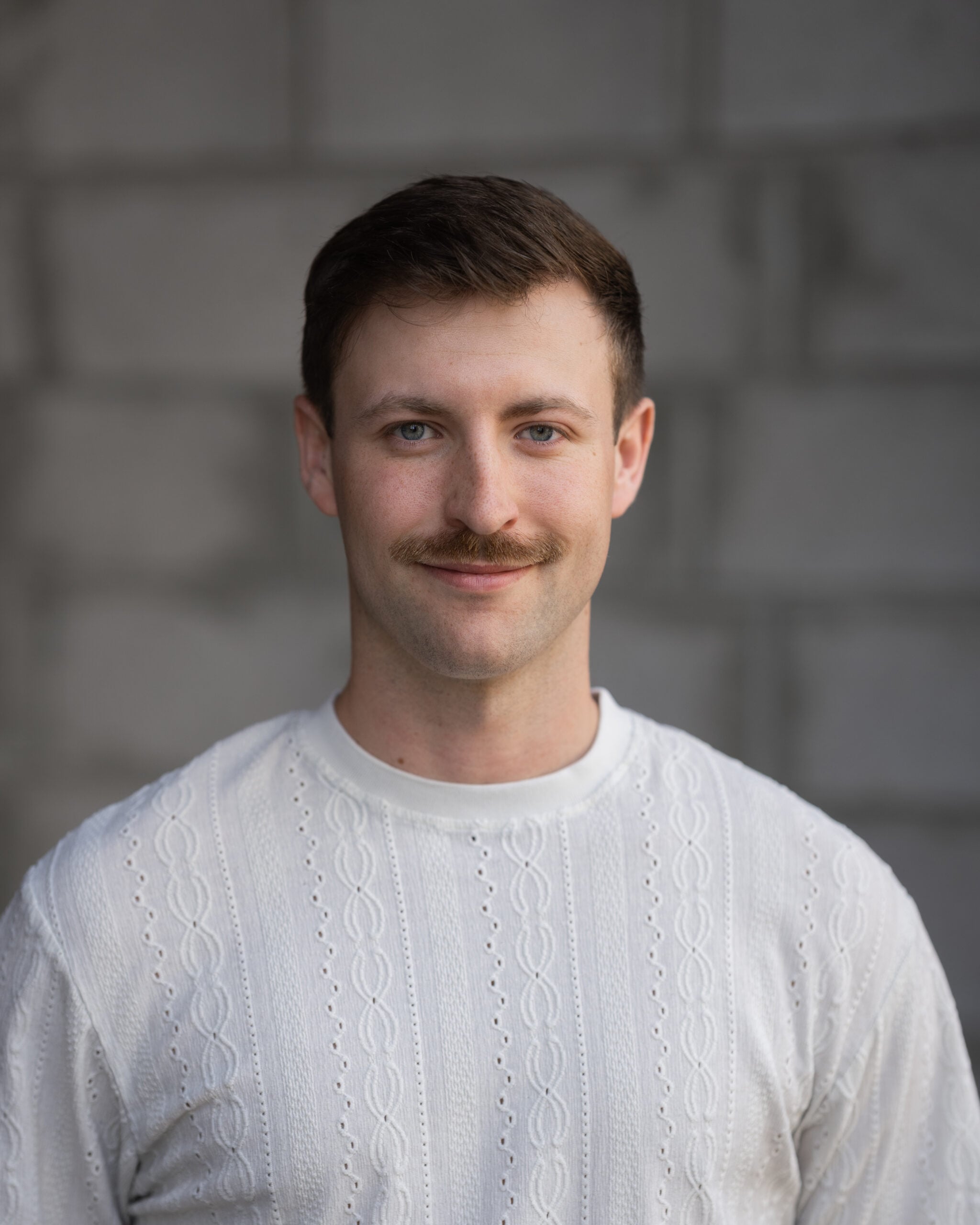 Jacob DeBoer, with short brown hair and a mustache, wears a white sweater and smiles slightly at the camera against a gray brick wall.