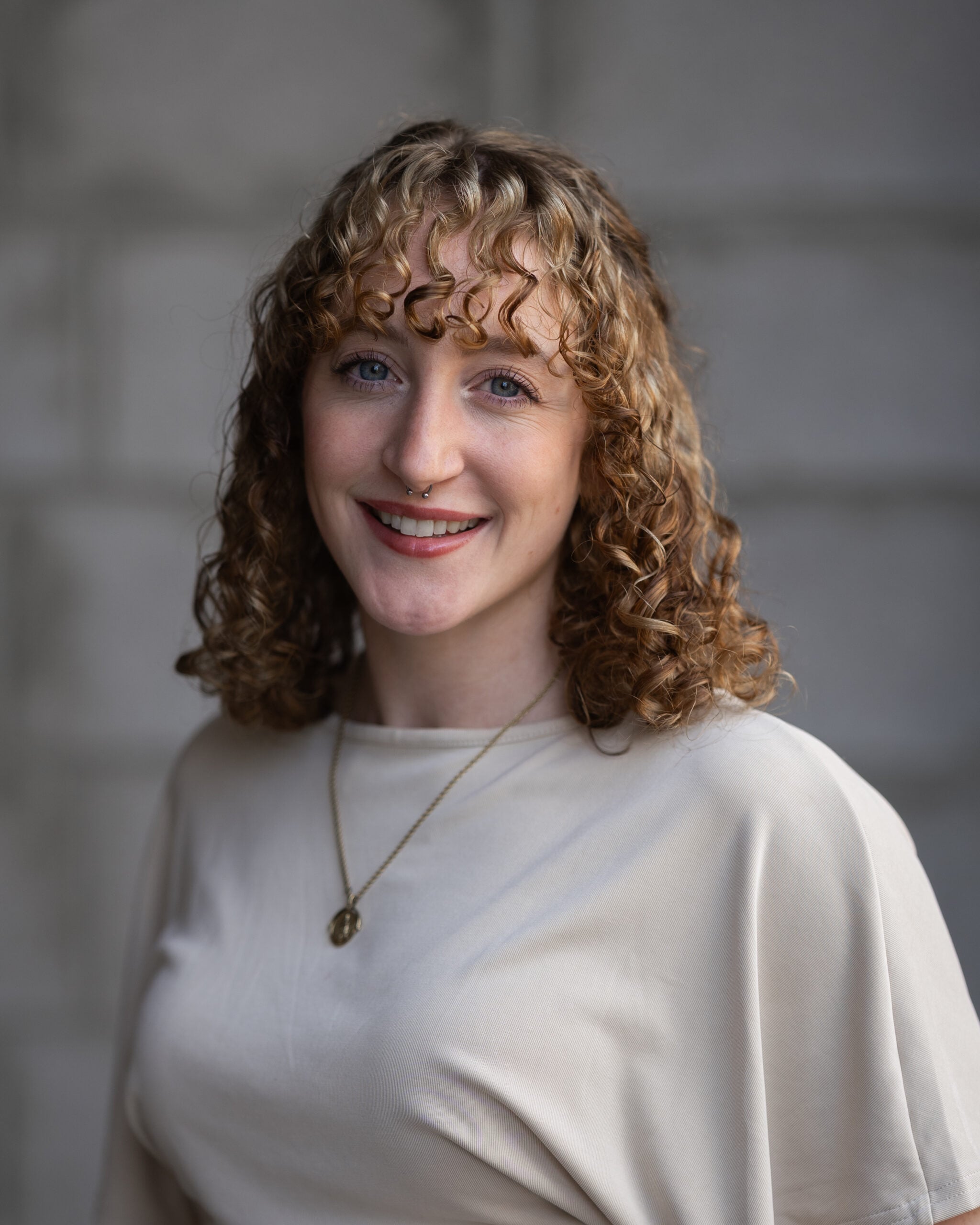 Smiling woman with curly light brown hair and blue eyes, in a cream top and pendant necklace. Blurred gray stone background.