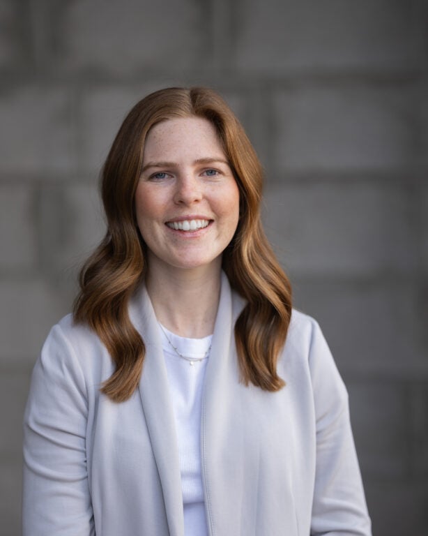 Megan Talbot, with long red hair, wears a light gray blazer and white top, smiling confidently before a gray textured backdrop.