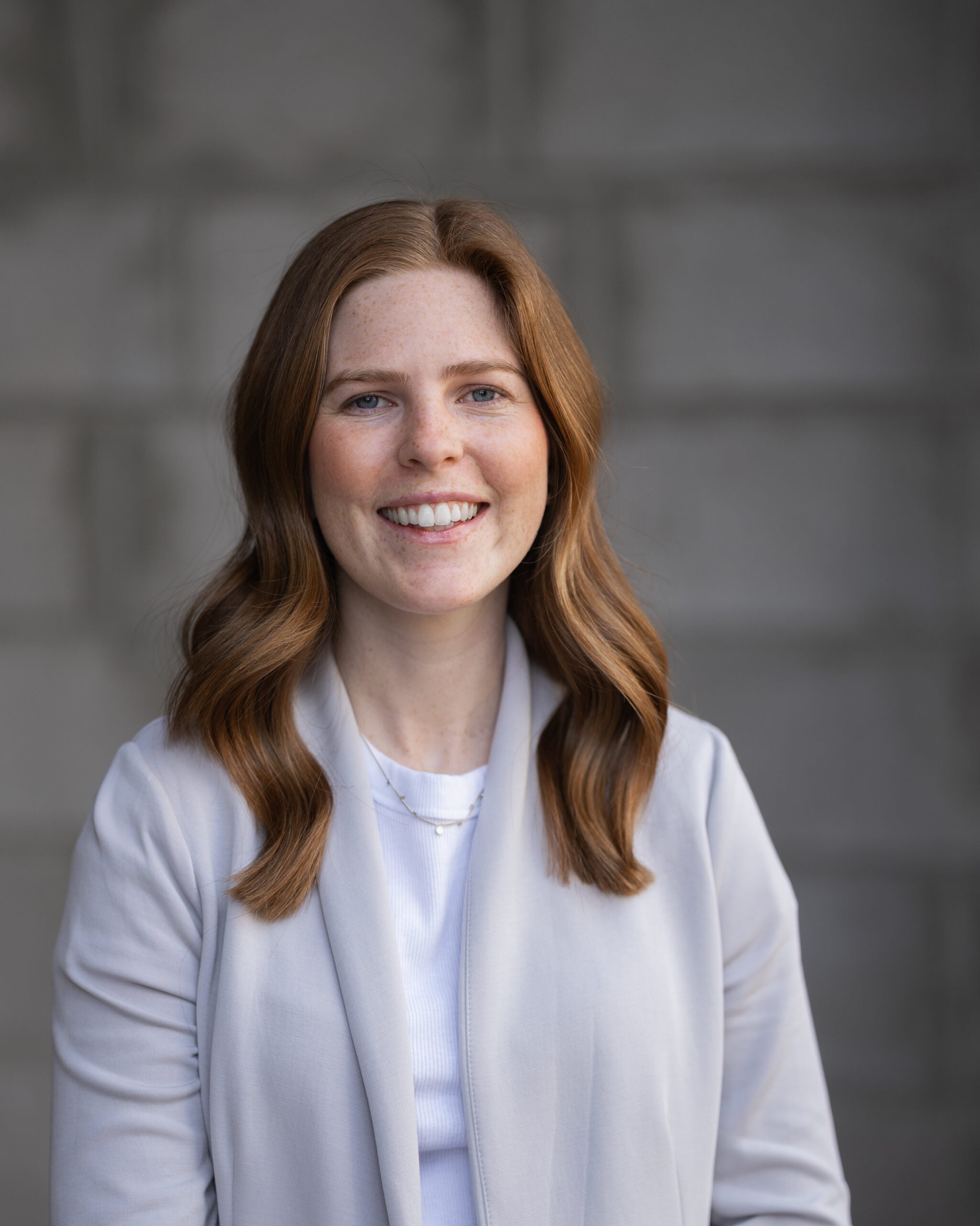 Megan Talbot, with long red hair, wears a light gray blazer and white top, smiling confidently before a gray textured backdrop.