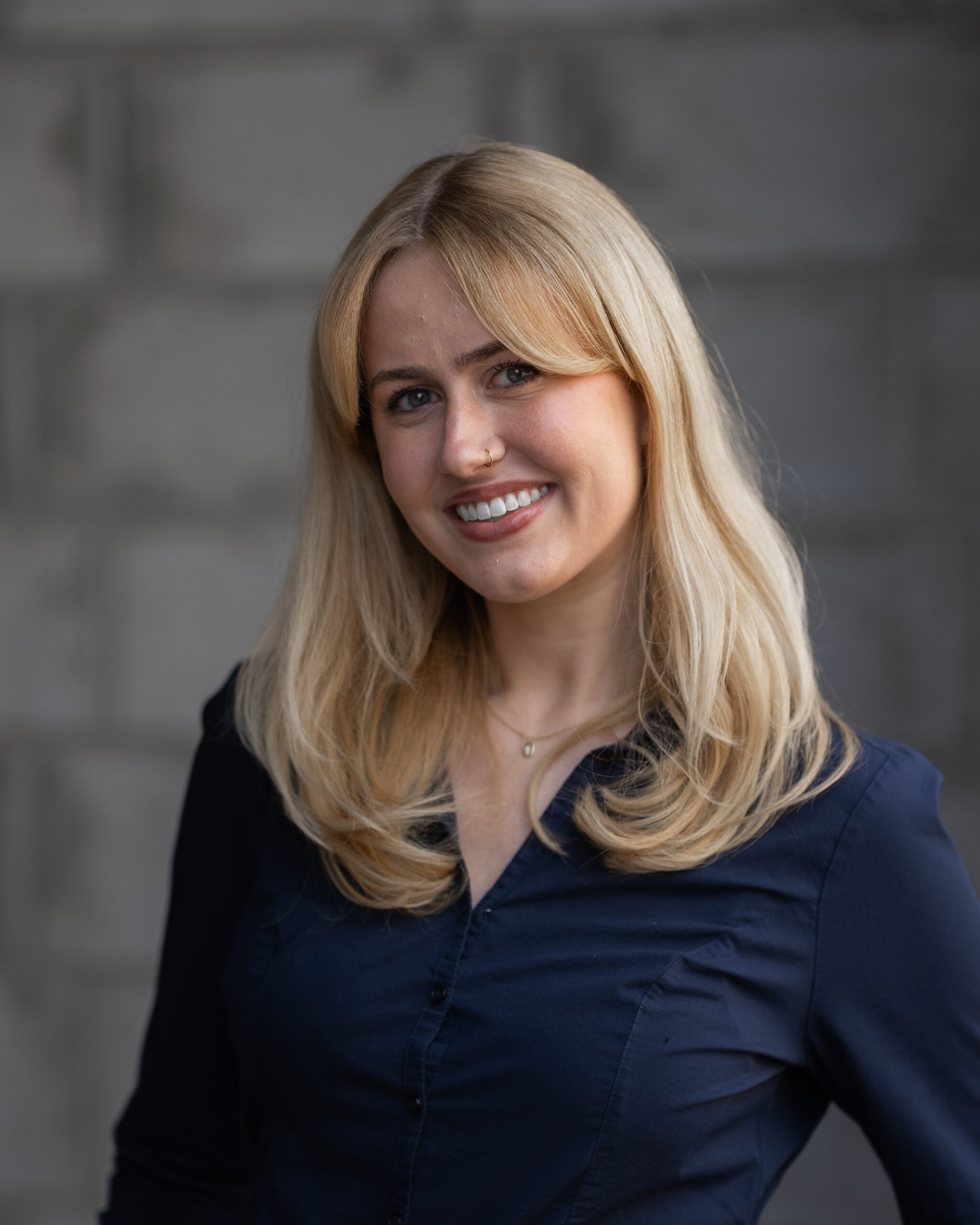 Professional woman with long blonde hair in a navy shirt, smiling confidently; blurred gray concrete blocks background.