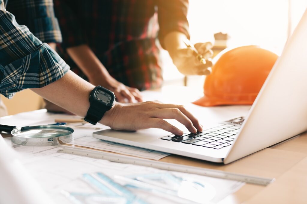 Image depicts a professional construction setting: Two individuals are engaged at a desk, one reviewing documents on a laptop while the other highlights key details with a pen. A magnifying glass, ruler, and orange safety helmet are also present, emphasizing precision and safety in engineering or construction work.