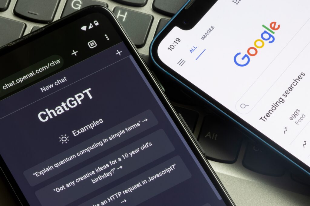 On a laptop keyboard, two smartphones are displayed: one features the ChatGPT homepage with vivid examples of AI-driven prompts, while the other displays Google's search homepage with trending topics.