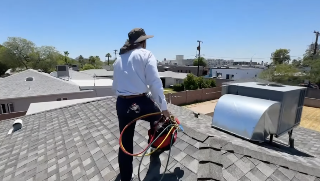 In 2025, an HVAC technician, equipped with a wide-brimmed hat and vibrant hoses, confidently navigates a shingled roof. They walk past a large metal vent against the backdrop of sunny residences, surrounded by trees and power lines under a clear sky.