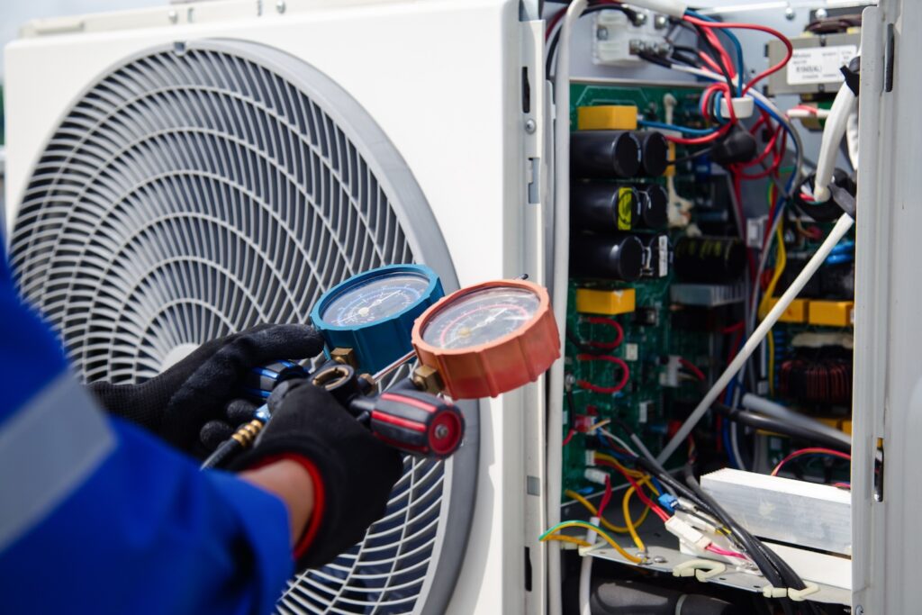 A technician in a blue uniform and black gloves expertly inspects an air conditioning unit with a manifold gauge set, highlighting the internal components and wiring. This essential home service guarantees optimal performance—an ideal blog topic to generate more leads.