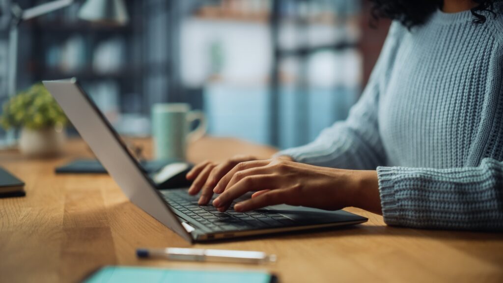 On Hook Agency's website, a focused individual in a light blue sweater types on a laptop at a wooden table, diligently generating ideas for an AI project. In the foreground, a pen and notebook lie ready for notes, while a mug and blurred bookshelf create an inspiring backdrop.