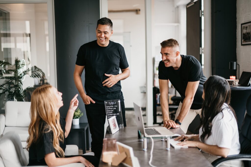 In an office setting at Hook Agency, four individuals engage in a dynamic conversation around a laptop on the table. With two standing and two seated, the scene reflects a collaborative atmosphere, underscoring the value of diverse perspectives.