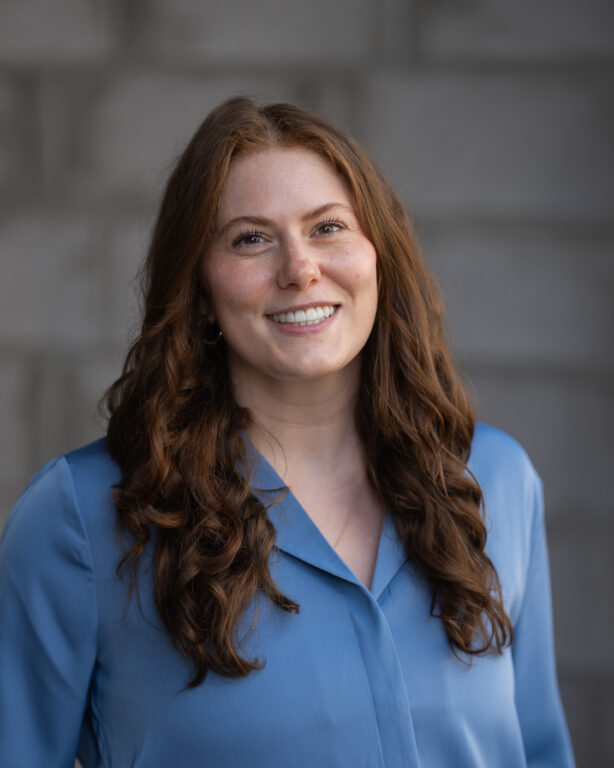 Madelynn Wald, Hook Agency team member, smiles at the camera in a light blue blouse, in front of a gray brick background.
