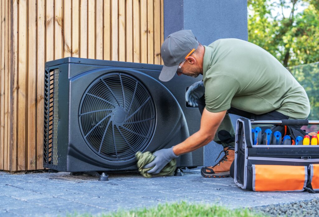 A certified technician in a green uniform and gloves cleans an outdoor heat pump unit—critical HVAC equipment—with a cloth, while a tool bag with essential tools is positioned nearby.