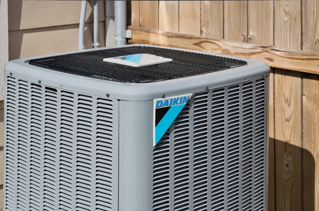 A top-rated gray Daikin outdoor air conditioning unit is professionally installed beside a wooden fence and the home's exterior wall, positioned for optimal performance and reliability.