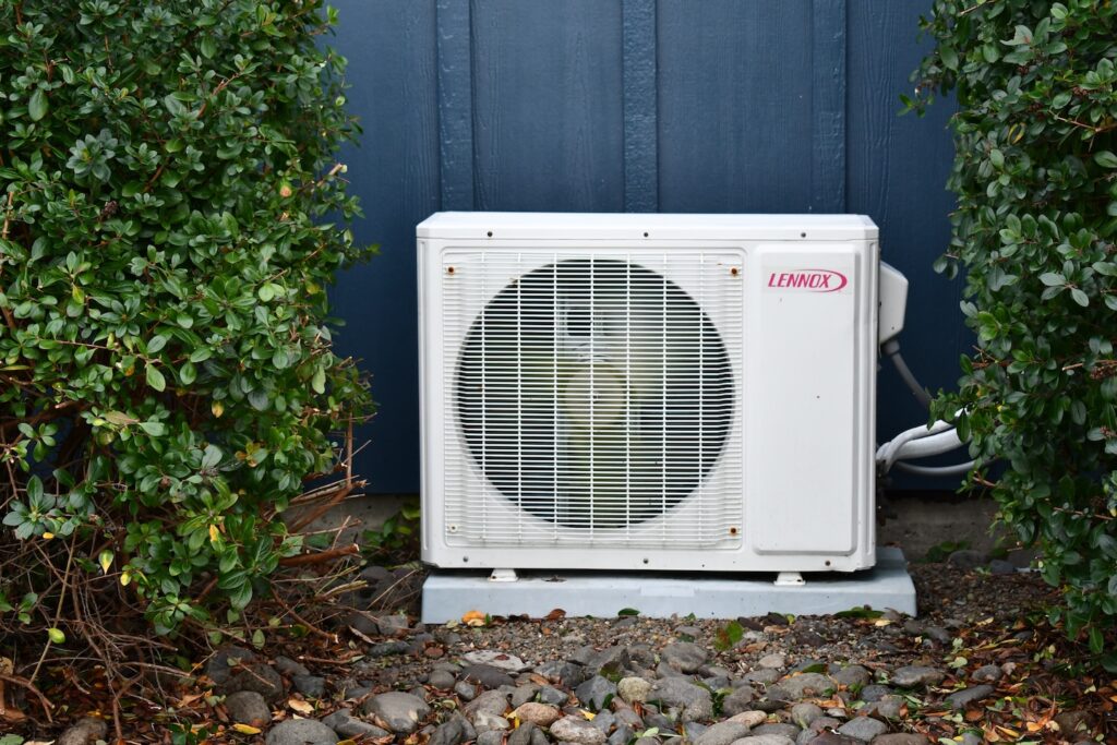 A white Lennox air conditioning unit, part of the building’s HVAC system, is installed on a concrete pad and positioned in front of a dark blue wall, surrounded by green bushes and decorative rocks.