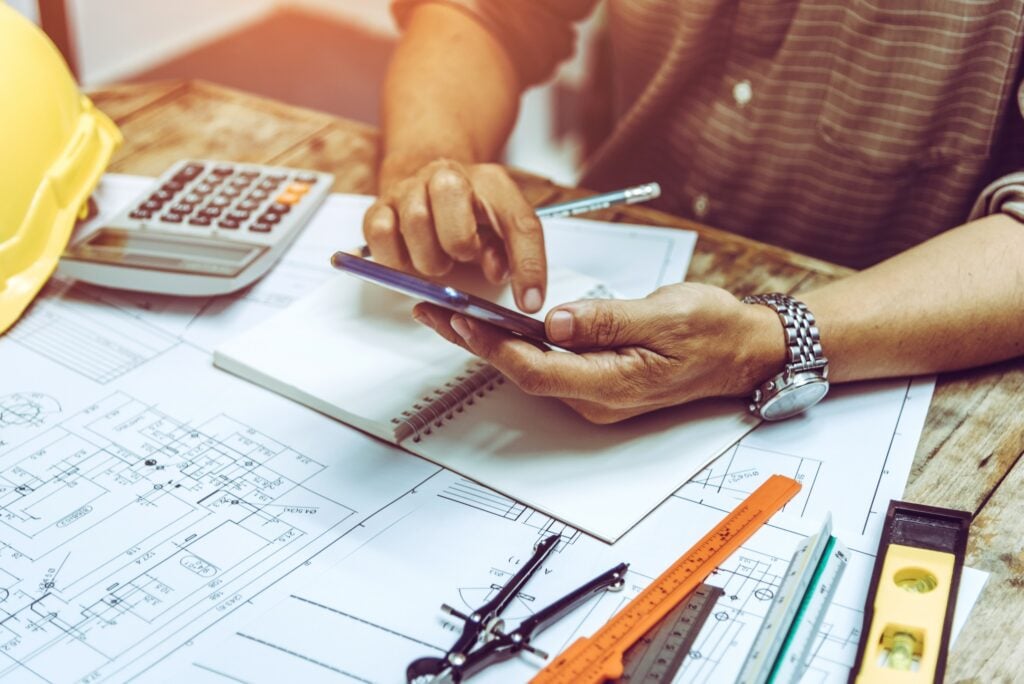 An industry leader in efficient home services, utilizes cutting-edge technology at a workstation adorned with technical drawings, a notebook, drafting tools, a calculator, and safety helmet.