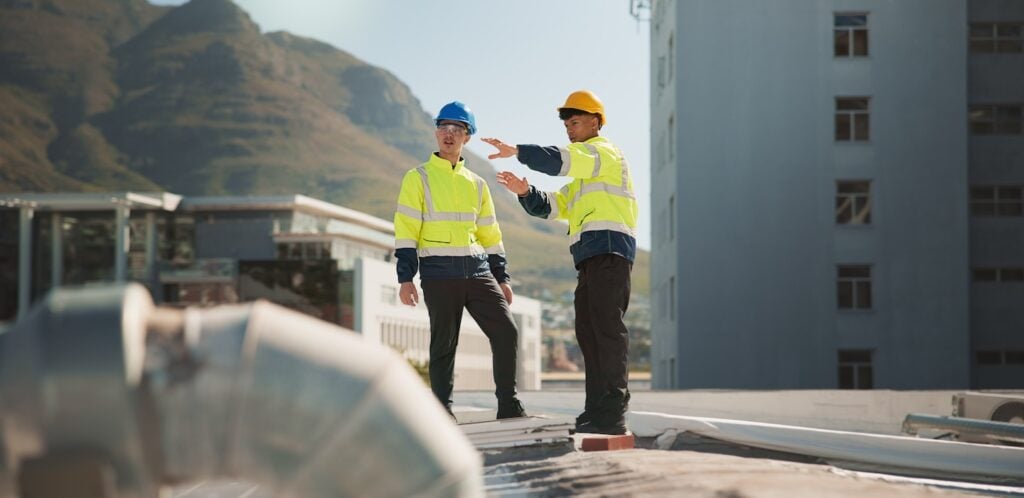 Two construction workers in high-visibility jackets and hard hats assess HVAC solutions on a rooftop, with city buildings and mountains in the background.
