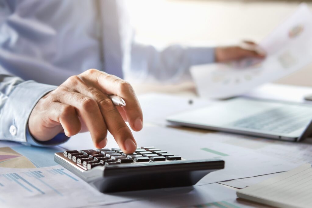 In a brightly lit office, a professional meticulously calculates figures from an essential business valuation guide. Amid a desk filled with paperwork and a laptop, they hold an important document, symbolizing the balance of precision and analysis in this bustling work environment.