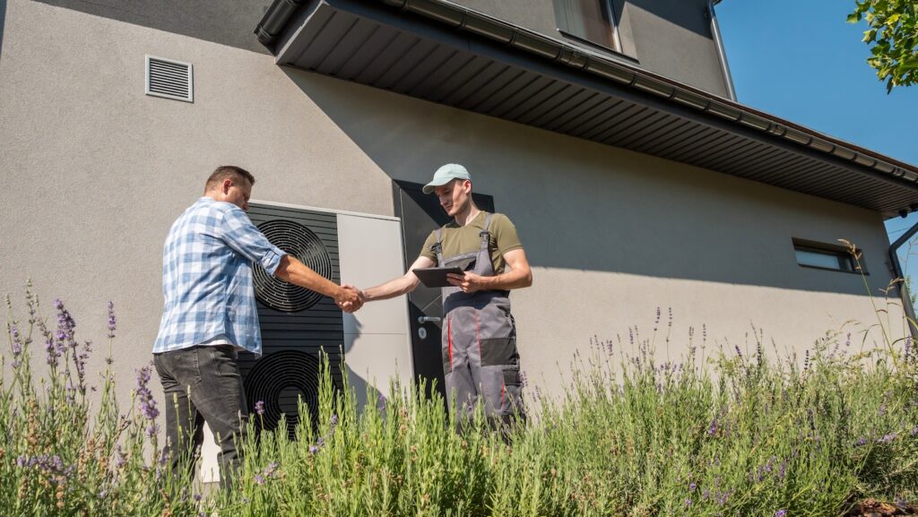 An HVAC technician, equipped with a tablet, shakes hands with a homeowner outside a modern residence beside an outdoor heat pump. Tall grass and plants frame the scene.