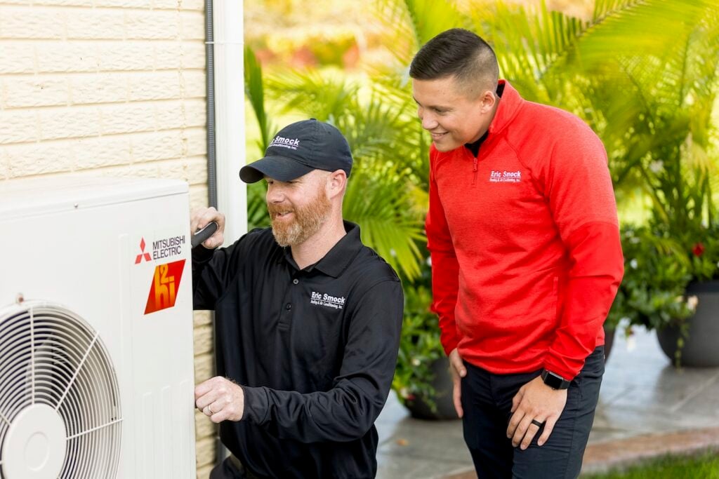 Two uniformed HVAC technicians service a Mitsubishi Electric outdoor unit—one adjusts the system while the other observes. Lush greenery frames the worksite.
