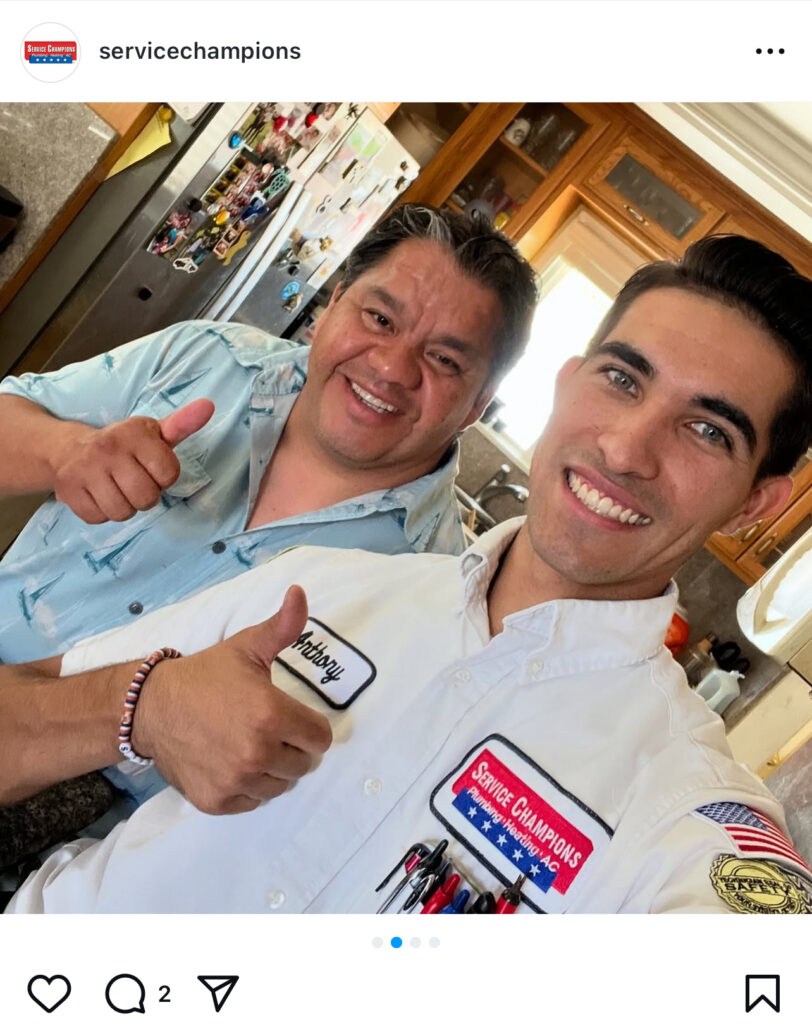 Two men stand smiling and giving a thumbs up in a modern kitchen—one in a blue shirt, the other in a Service Champions HVAC uniform with an American flag patch and name tag. Refrigerator and cabinets frame the scene. Ideal for Hook Agency’s 2025 social media content showcasing satisfied customers and professional service.