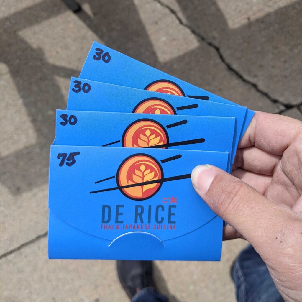 A hand displays four "De Rice Thai & Japanese Cuisine" gift cards—ideal for employee appreciation. Three cards are marked "$30" and one is marked "$75." The background includes pavement and a shoe.