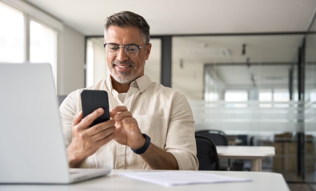 A smiling man with glasses manages home services social media from a modern office, seamlessly using his smartphone and laptop. A sheet of paper rests on the desk in front of him.