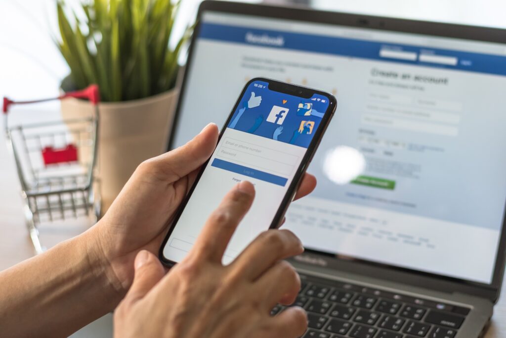 A smartphone displays the Facebook login screen, with a laptop in the background also open to Facebook’s login and sign-up page—representing effective management of home services Facebook ads. A small shopping cart and plant complete the workspace setting.