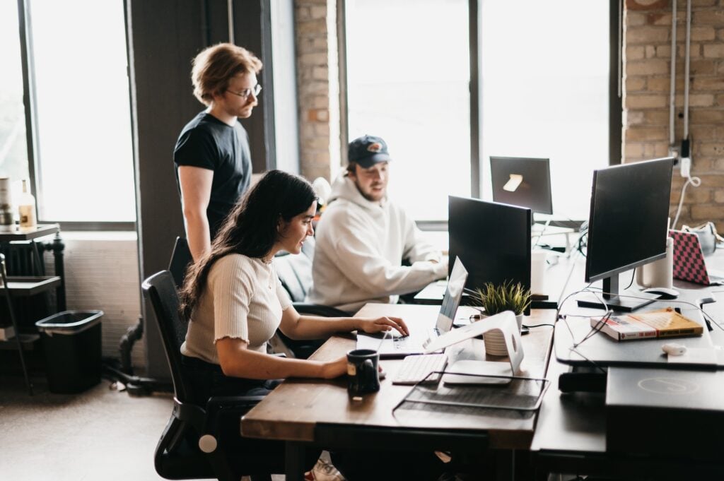 Three team members collaborate at a modern office desk, evaluating the choice between in-house marketing and partnering with an agency. One stands while two are seated—all focused on their screens as natural light fills the workspace through large windows.